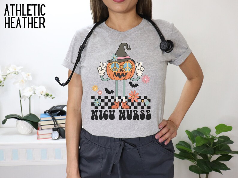 Puede incluir: Una camiseta gris jaspeada con un gr&aacute;fico de una calabaza con un sombrero de bruja y gafas de sol. La calabaza est&aacute; haciendo el signo de la paz y est&aacute; de pie sobre un patr&oacute;n a cuadros. El texto "NICU NURSE" est&aacute; impreso debajo de la calabaza.