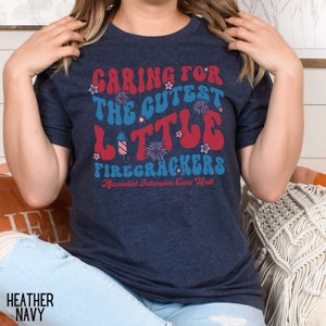 May include: A navy blue t-shirt with a red, white, and blue graphic that says "Caring for the Cutest Little Firecrackers" and "Neonatal Intensive Care Unit".