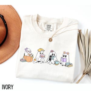 May include: Ivory-colored Comfort Colors t-shirt featuring a graphic of four cartoon ghosts reading books. One ghost is sitting on a pumpkin. The shirt has a round neckline and short sleeves. A gold necklace is draped on the shirt.