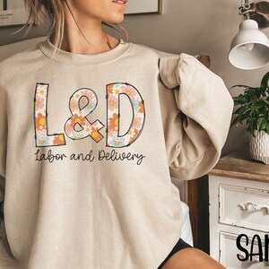 May include: Sand-colored sweatshirt with the letters "L&D" in a floral design, with the words "Labor and Delivery" underneath. The sweatshirt is a neutral color and has long sleeves.