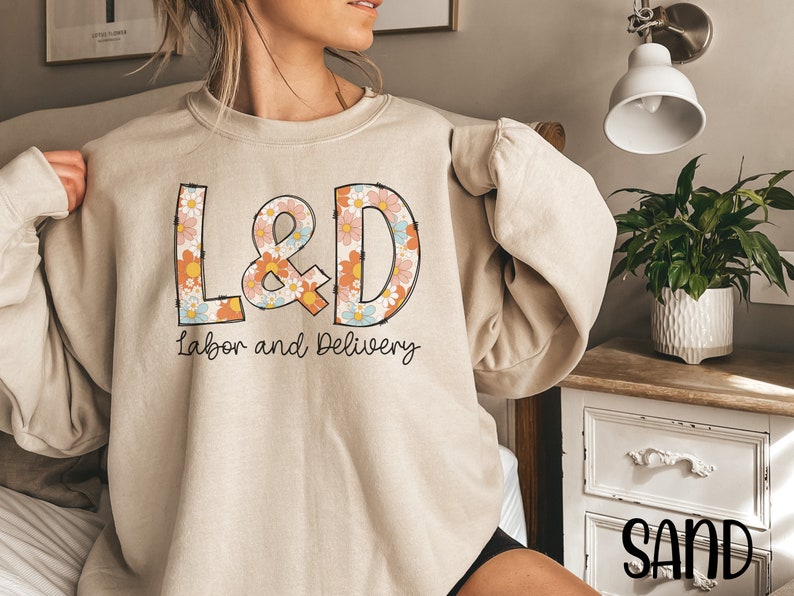 May include: Sand-colored sweatshirt with the letters "L&D" in a floral design, with the words "Labor and Delivery" underneath. The sweatshirt is a neutral color and has long sleeves.