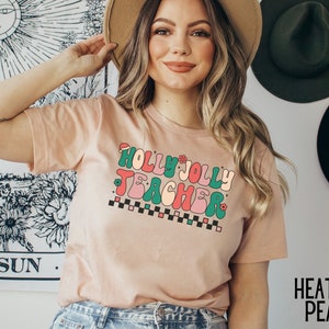 May include: A light pink t-shirt with a retro-style graphic that says "Holly Jolly Teacher" in green, pink, and red letters with a checkered pattern underneath.