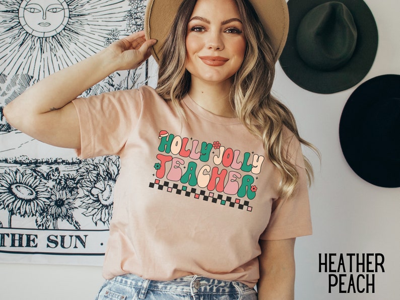May include: A light pink t-shirt with a retro-style graphic that says "Holly Jolly Teacher" in green, pink, and red letters with a checkered pattern underneath.