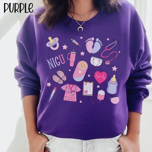 May include: Purple sweatshirt with a variety of colorful illustrations, including a baby in a swaddle, a stethoscope, a pacifier, a syringe, a baby bottle, a heart monitor, a thermometer, a pair of shoes, a cup, a diaper, a medical chart, a blood bag, and a face mask. The text "NICU" is printed on the sweatshirt.