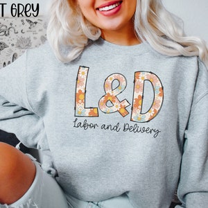May include: Sport grey sweatshirt with the text "L&D Labor and Delivery" in a retro floral design. The sweatshirt is light grey and has long sleeves. The text "Sport Grey" is at the top left.