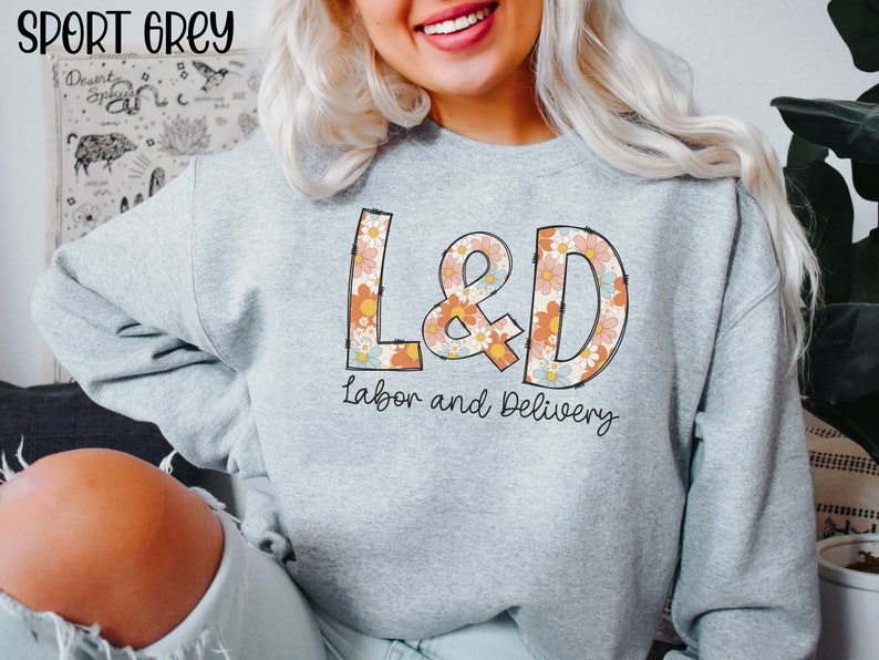 May include: Sport grey sweatshirt with the text "L&D Labor and Delivery" in a retro floral design. The sweatshirt is light grey and has long sleeves. The text "Sport Grey" is at the top left.