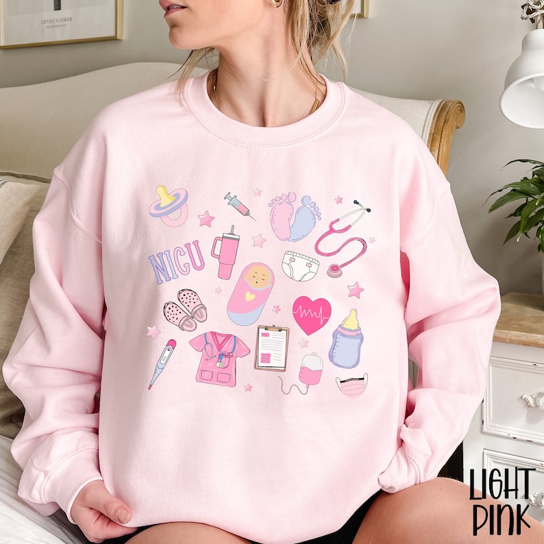 May include: Pink sweatshirt with a design of various medical symbols and the word "NICU" in white. The symbols include a stethoscope, a baby bottle, a pacifier, a syringe, a heart monitor, a thermometer, a baby footprint, a diaper, a baby in a crib, and a nurse's uniform.
