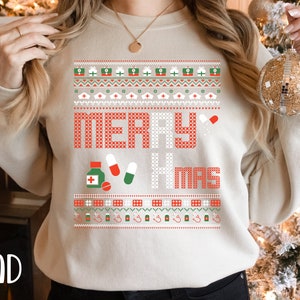 K&ouml;nnte beinhalten: Ein beiger Pullover mit einem roten und gr&uuml;nen Weihnachtsmotiv. Das Design zeigt die Worte "Merry Xmas" und verschiedene medizinische Symbole, darunter Pillen, eine Flasche Medizin und ein Stethoskop.