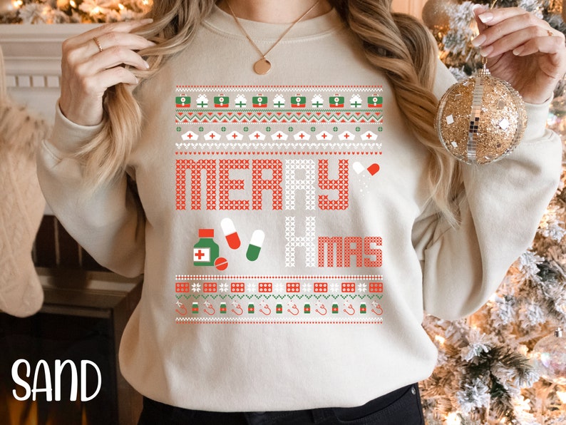 K&ouml;nnte beinhalten: Ein beiger Pullover mit einem roten und gr&uuml;nen Weihnachtsmotiv. Das Design zeigt die Worte "Merry Xmas" und verschiedene medizinische Symbole, darunter Pillen, eine Flasche Medizin und ein Stethoskop.