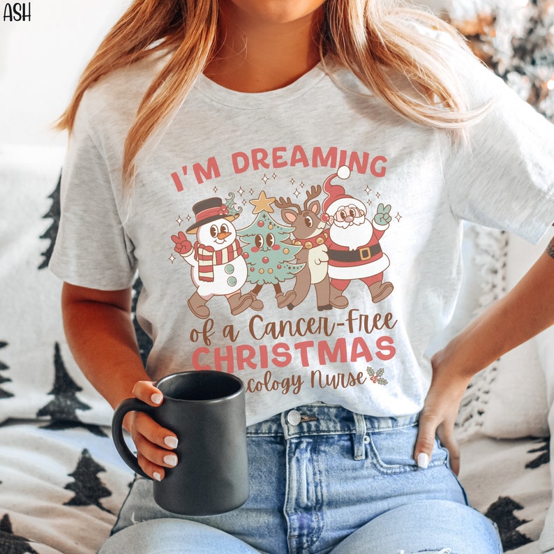 May include: A white t-shirt with the text "I'm Dreaming of a Cancer-Free Christmas Oncology Nurse" and a cartoon image of Santa Claus, a snowman, a reindeer, and a Christmas tree.
