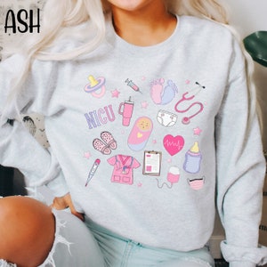 May include: A light gray sweatshirt with a pink and blue graphic design featuring the text "NICU" and various medical symbols, including a stethoscope, a syringe, a baby bottle, a pacifier, a thermometer, a heart rate monitor, and a baby footprint.