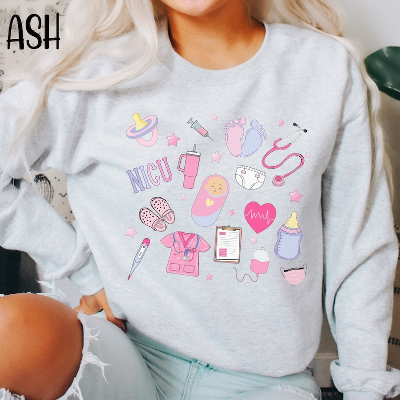 May include: A light gray sweatshirt with a pink and blue graphic design featuring the text "NICU" and various medical symbols, including a stethoscope, a syringe, a baby bottle, a pacifier, a thermometer, a heart rate monitor, and a baby footprint.
