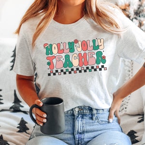 May include: A white t-shirt with the text "Holly Jolly Teacher" in a retro, colorful font. The text is surrounded by small red and green flowers and a black and white checkered pattern.