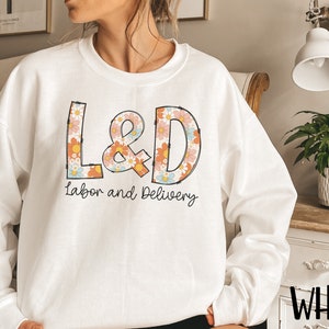 May include: White sweatshirt with the letters "L&D" in a floral design, with the words "Labor and Delivery" underneath. The sweatshirt is a crew neck style.