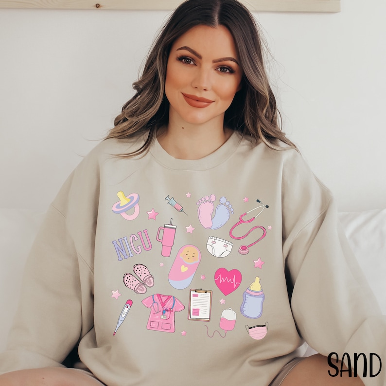 May include: A light beige sweatshirt with a graphic design featuring a variety of baby and medical icons, including a pacifier, syringe, baby footprints, stethoscope, a cup with the word "NICU", a baby doll, a diaper, a heart with a heartbeat line, a baby bottle, a thermometer, a nurse's uniform, a clipboard, a blood pressure cuff, and a face mask.