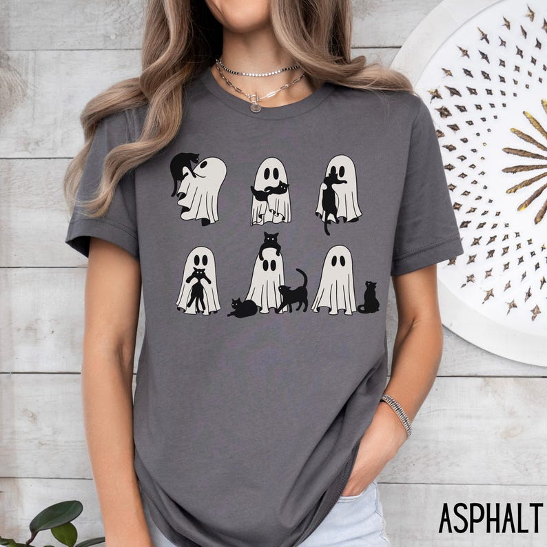 Puede incluir: Una camiseta gris con un gr&aacute;fico de gatos negros asom&aacute;ndose de fantasmas blancos. El texto "ASPHALT" es visible en la esquina inferior derecha de la imagen.
