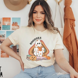 May include: A woman wearing a pale yellow t-shirt with a ghost reading a book and the text "BOOOKS" printed on it. The ghost is sitting on a stack of books with a pumpkin in front of it. The ghost is surrounded by a rainbow.