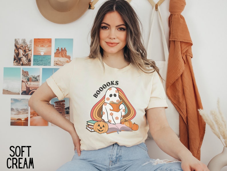 May include: A woman wearing a pale yellow t-shirt with a ghost reading a book and the text "BOOOKS" printed on it. The ghost is sitting on a stack of books with a pumpkin in front of it. The ghost is surrounded by a rainbow.