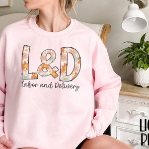 May include: Light pink sweatshirt with the text "L&D Labor and Delivery" in a retro floral design. The sweatshirt is a crew neck style and has long sleeves. The text "Light Pink" is in the bottom right corner.