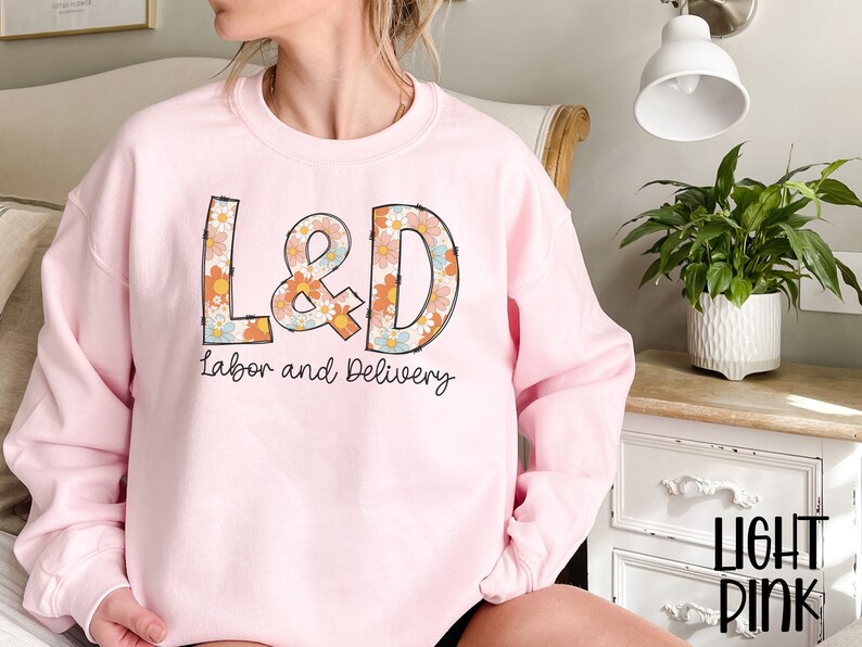May include: Light pink sweatshirt with the text "L&D Labor and Delivery" in a retro floral design. The sweatshirt is a crew neck style and has long sleeves. The text "Light Pink" is in the bottom right corner.