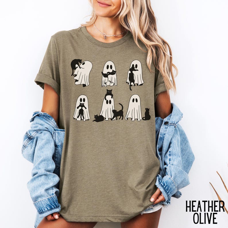 Puede incluir: Una camiseta de color oliva jaspeado con un gr&aacute;fico de fantasmas y gatos. Los fantasmas son blancos y los gatos son negros. El texto "HEATHER OLIVE" est&aacute; impreso en la esquina inferior derecha de la imagen.