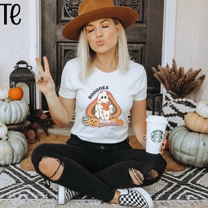 May include: White t-shirt featuring a ghost holding a book with the text "BOOOKS" in rainbow letters. The ghost is wearing a pumpkin hat and sitting on a pile of books. The shirt is being worn by a person sitting on a rug with pumpkins and autumnal decor around them.