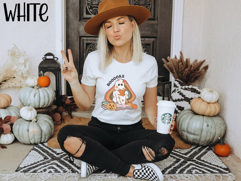 May include: White t-shirt featuring a ghost holding a book with the text "BOOOKS" in rainbow letters. The ghost is wearing a pumpkin hat and sitting on a pile of books. The shirt is being worn by a person sitting on a rug with pumpkins and autumnal decor around them.