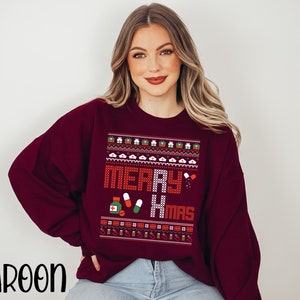 K&ouml;nnte beinhalten: Ein weinroter Weihnachtspullover mit einem pixeligen Design in Wei&szlig;, Rot und Gr&uuml;n, auf dem "Merry Xmas" steht. Das Design enth&auml;lt ein Herz, Pillen und eine Erste-Hilfe-Box.