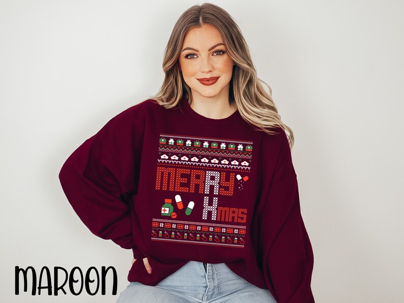 K&ouml;nnte beinhalten: Ein weinroter Weihnachtspullover mit einem pixeligen Design in Wei&szlig;, Rot und Gr&uuml;n, auf dem "Merry Xmas" steht. Das Design enth&auml;lt ein Herz, Pillen und eine Erste-Hilfe-Box.