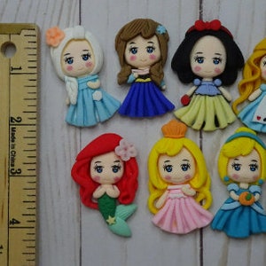 May include: A collection of handcrafted princess figurines, each with unique hairstyles and colorful dresses. The figurines are approximately 1.5 inches tall and are arranged on a light wood surface. A wooden ruler is placed next to the figurines.