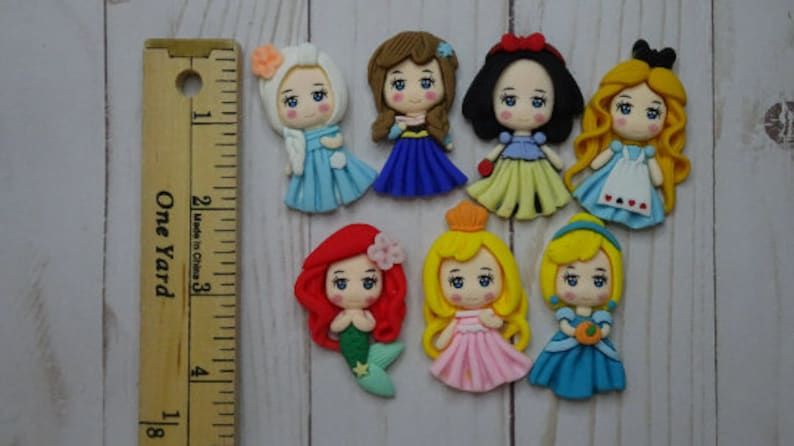 May include: A collection of handcrafted princess figurines, each with unique hairstyles and colorful dresses. The figurines are approximately 1.5 inches tall and are arranged on a light wood surface. A wooden ruler is placed next to the figurines.