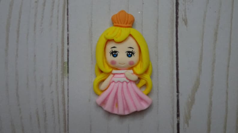May include: A princess figurine with a yellow crown and long, flowing yellow hair. She wears a pink dress and has blue eyes and rosy cheeks. The figurine is likely made of polymer clay.