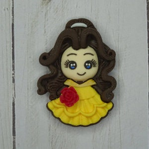 May include: A decorative figurine of a cartoon princess with brown hair, blue eyes, and a yellow dress. A red rose is on the front of the dress. The figurine is on a white wooden surface.