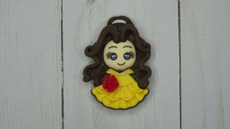 May include: A decorative figurine of a cartoon princess with brown hair, blue eyes, and a yellow dress. A red rose is on the front of the dress. The figurine is on a white wooden surface.
