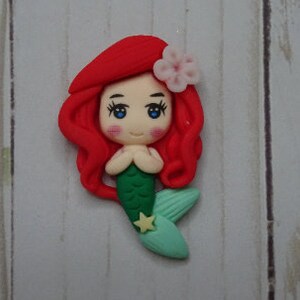 May include: A handcrafted mermaid figurine with red hair, blue eyes, and a green tail. The mermaid has a pink flower in her hair and a yellow star on her tail. The figurine is made of clay.