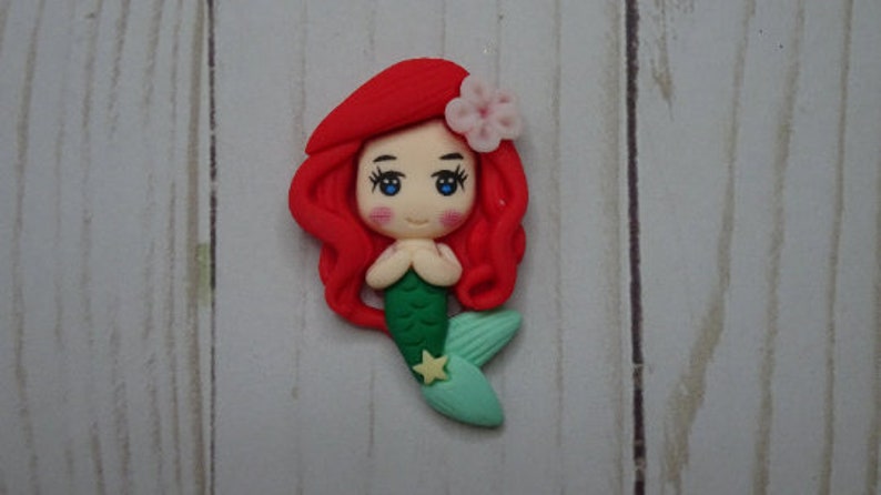 May include: A handcrafted mermaid figurine with red hair, blue eyes, and a green tail. The mermaid has a pink flower in her hair and a yellow star on her tail. The figurine is made of clay.