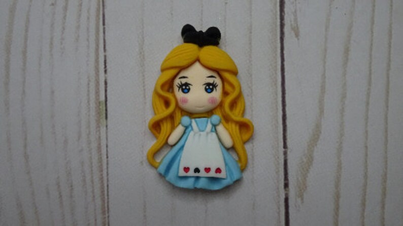 May include: A handcrafted figurine of Alice from Alice in Wonderland. The figurine features a light blue dress, white apron with heart details, and long, flowing yellow hair. A black bow sits atop her head. The figurine is set against a light wood grain background.