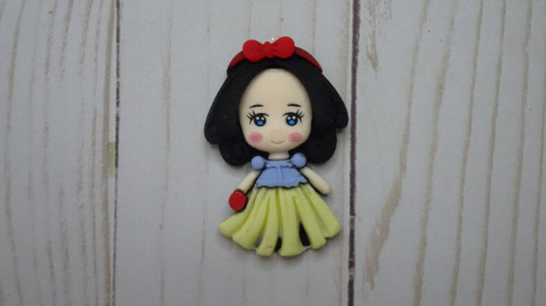 May include: A handcrafted Snow White figurine with black hair, blue eyes, and a red bow. The doll wears a blue top, a yellow skirt, and red gloves. The figurine is a charming representation of the classic fairytale character.