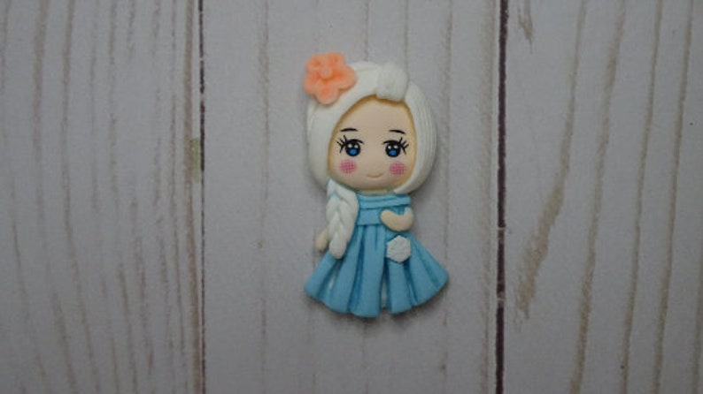 May include: A handcrafted figurine of a cartoon girl with white hair and a blue dress. The figurine has a peach-colored flower in her hair and a white braid. The girl has blue eyes and rosy cheeks. The figurine is on a light wood grain surface.