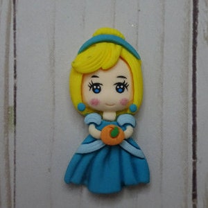May include: A figurine of a princess with yellow hair, blue eyes, and a blue dress, holding an orange pumpkin. The princess has rosy cheeks and is wearing a blue headband and earrings. The background is a light wood grain.