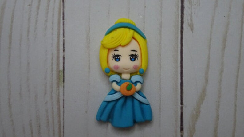 May include: A figurine of a princess with yellow hair, blue eyes, and a blue dress, holding an orange pumpkin. The princess has rosy cheeks and is wearing a blue headband and earrings. The background is a light wood grain.