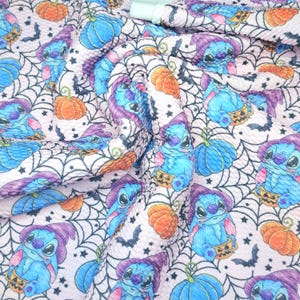 May include: A light pink fabric featuring a repeating pattern of blue alien characters wearing purple witch hats, orange pumpkins, black spiderwebs, and small black bats. The material has a textured, slightly puckered surface throughout.