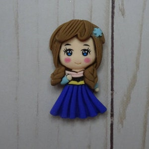 May include: A handcrafted figurine of a cartoon girl with brown hair in braids, blue eyes, and rosy cheeks. She wears a blue dress, a pink and black top, and a blue flower in her hair. The figurine is set against a light wood grain background.
