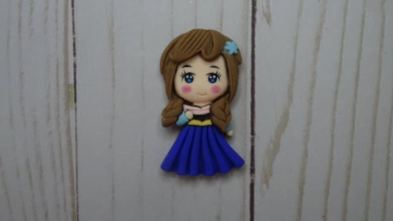 May include: A handcrafted figurine of a cartoon girl with brown hair in braids, blue eyes, and rosy cheeks. She wears a blue dress, a pink and black top, and a blue flower in her hair. The figurine is set against a light wood grain background.