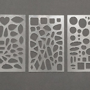 May include: Three silver metal stencils with various cut-out shapes, including hearts, teardrops, squares, and circles. The stencils are arranged side-by-side on a gray surface, providing a variety of design options for crafts or art projects.