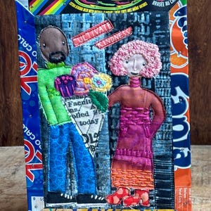 May include: A mixed-media textile art piece featuring two embroidered figures on a dark fabric background, framed by a colorful recycled beverage carton border. The text "CULTIVATE KINDNESS" is stitched above the figures, who hold a bouquet.