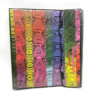 May include: A Trapper Keeper binder featuring a vertical striped pattern with swirling, marbled textures in vibrant colors including red, yellow, pink, green, blue, and purple. The front flap displays the text Trapper Keeper Design Series.