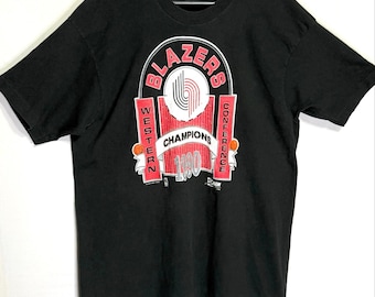 Puede incluir: Una camiseta negra de manga corta con un estampado gráfico que dice BLAZERS WESTERN CONFERENCE CHAMPIONS 1990. El diseño incluye un logotipo de baloncesto rojo y blanco, dos balones de baloncesto naranjas y texto en letras blancas y rojas.