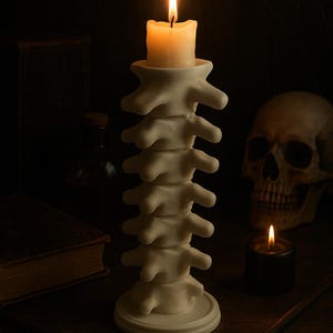 Pode incluir: Uma vela acesa está colocada em um castiçal em forma de coluna vertebral. O suporte esbranquiçado tem a forma de uma coluna vertebral humana. Uma caveira e outras velas estão no fundo, criando um tema gótico.
