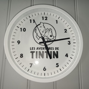 Può includere: Orologio da parete bianco con un'illustrazione in bianco e nero di Tintin e Milù. Il quadrante mostra numeri e lancette nere, con la scritta "LES AVENTURES DE TINTIN". L'orologio è rotondo.